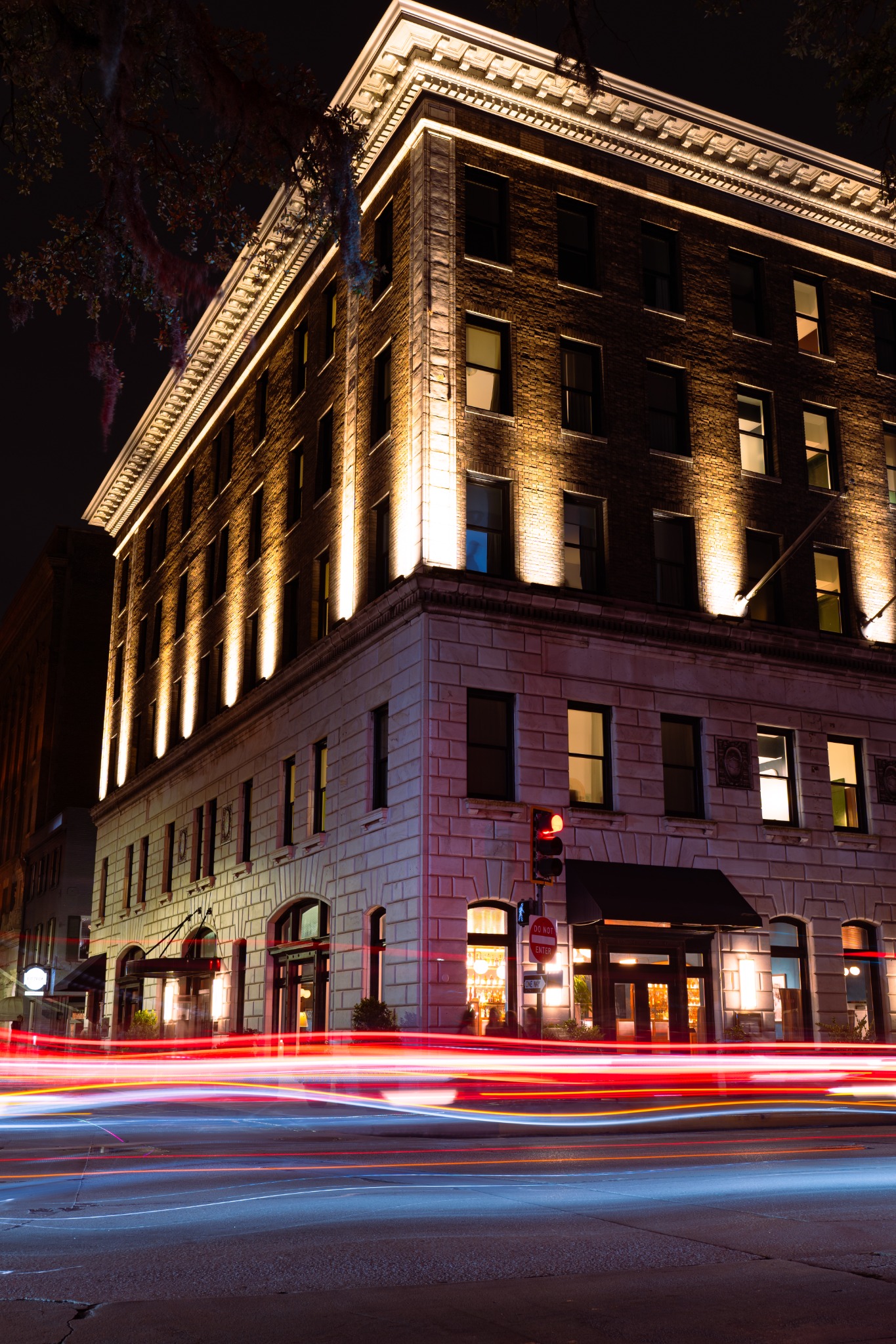 An illuminated brick building at night with blurred red and white traffic lights across the foreground